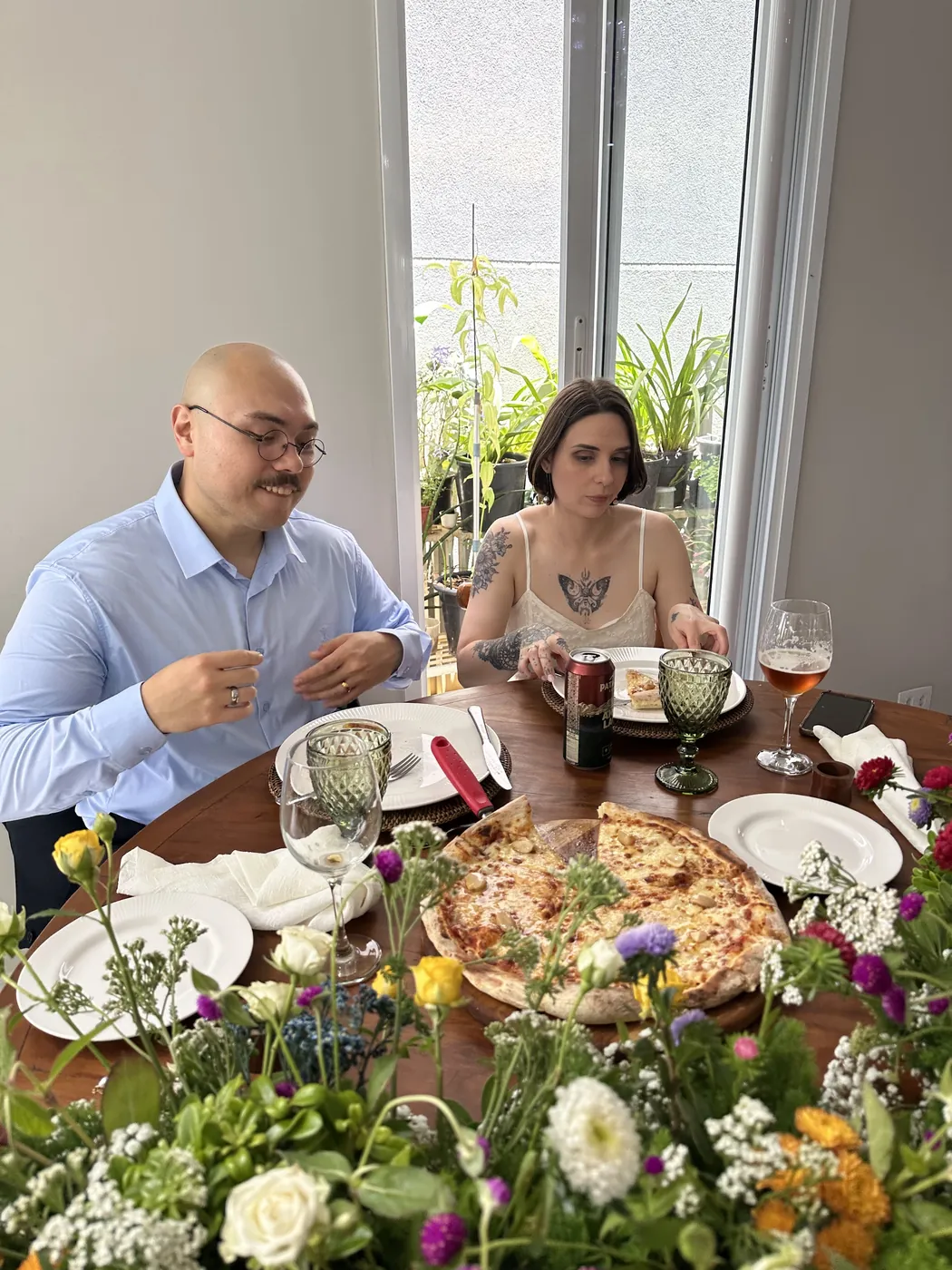 Os noivos à mesa decorada com flores, com a pizza servida, num casamento intimista